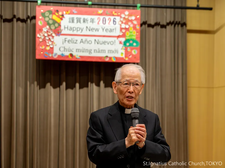 1/11(日)10:00- 主の洗礼の祝日 謹賀新年の赤い舞台看板の前で新年のあいさつをする髙祖神父